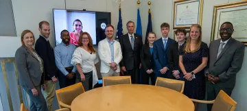 HS-POWER interns Dr. Nicole Fernandez Tejero (on tv screen), Mitch Harrison, Brooke Cochran, Paul Harris and Joseph Cruikshank standing with their mentors and HS-POWER Program Manager Greg Simmons (far right in gray suit) during a meet-and-greet with Dr. Dimitri Kusnezov, Under Secretary for Science and Technology (center, standing in front of flags).